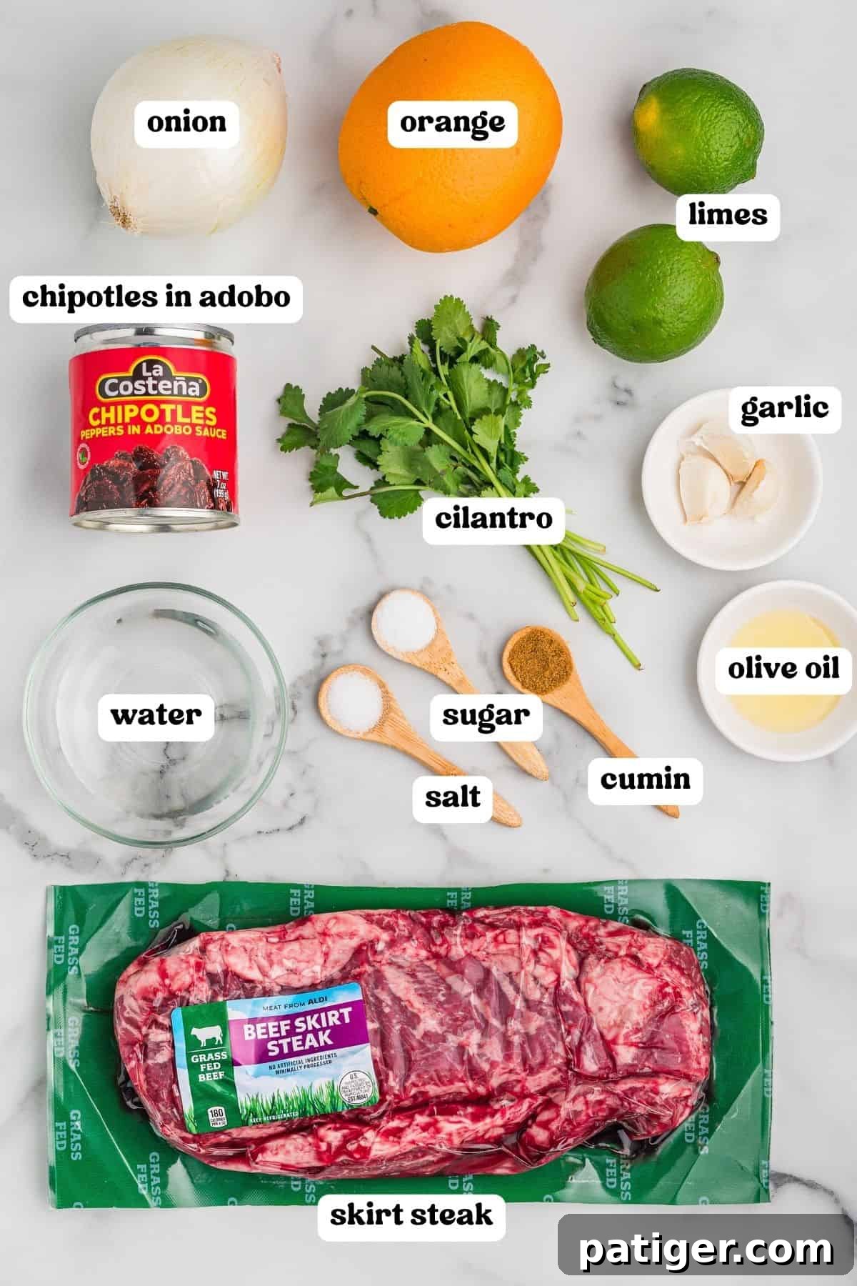 Juicy Slow Cooker Carne Asada Recipe 4 Labeled ingredients on a marble counter: onion, orange, limes, canned chipotles in adobo, cilantro, garlic, water, salt, sugar, cumin, olive oil, and skirt steak.