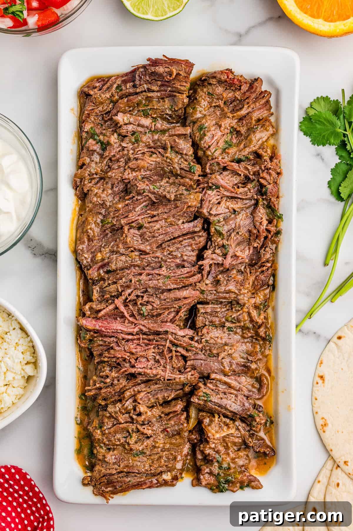 Juicy Slow Cooker Carne Asada Recipe 9 Sliced carne asada on a white platter surrounded by bowls of sour cream, cotija, pico de gallo, lime, and tortillas.
