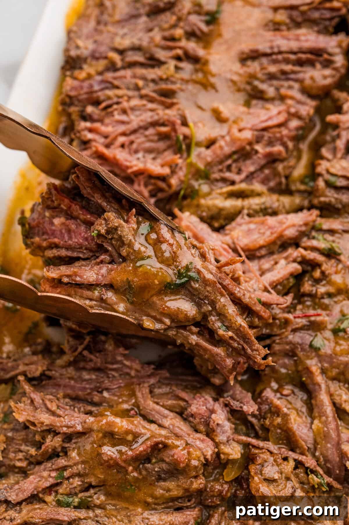 Juicy Slow Cooker Carne Asada Recipe 10 Tongs lifting a piece of saucy shredded beef from a white baking dish.