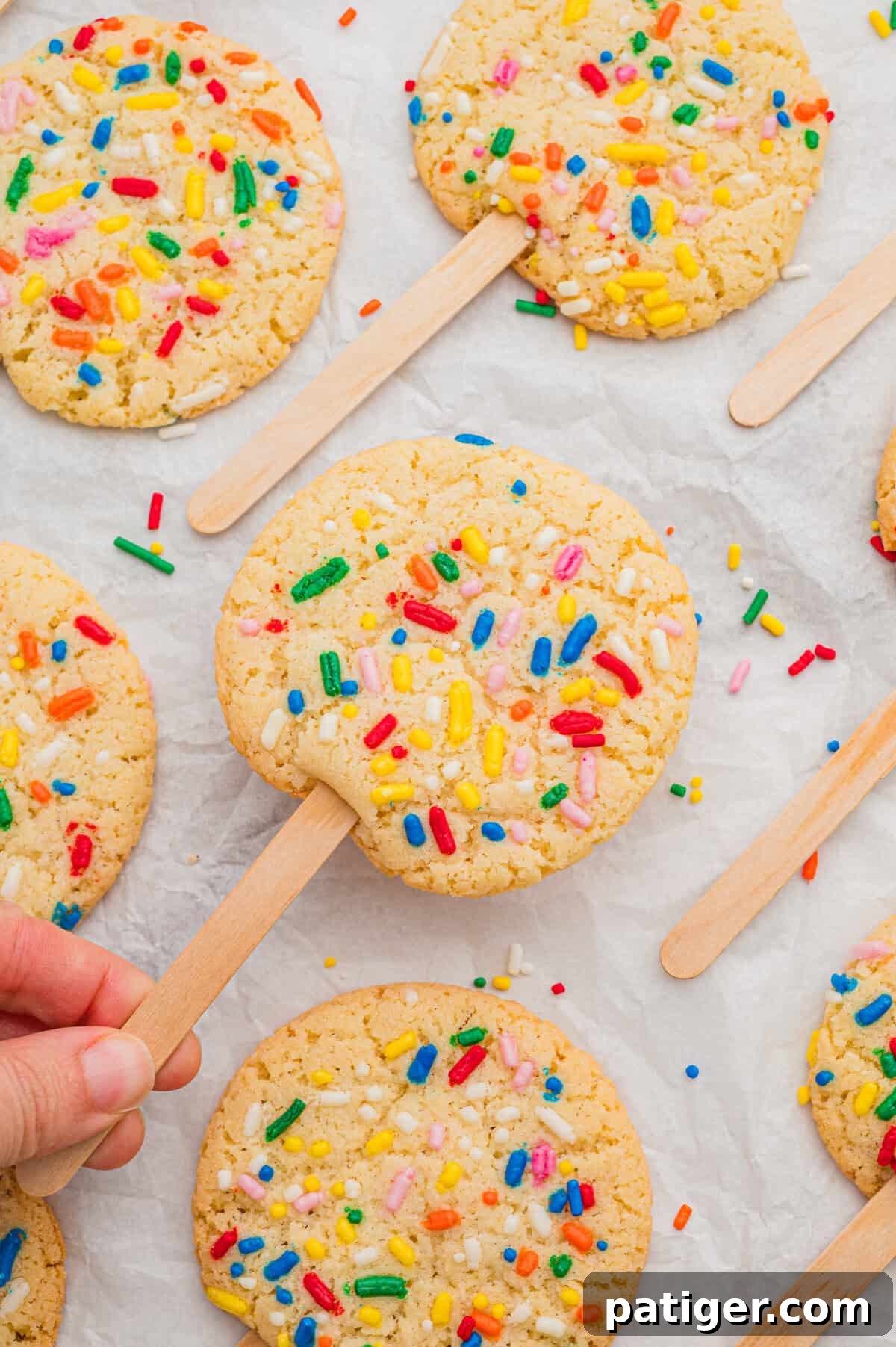 Cookie Pops: Make Handheld Cookies on a Stick 3 Cookies on a stick topped with rainbow sprinkles, one lifted by hand from a parchment-lined surface of matching cookies.