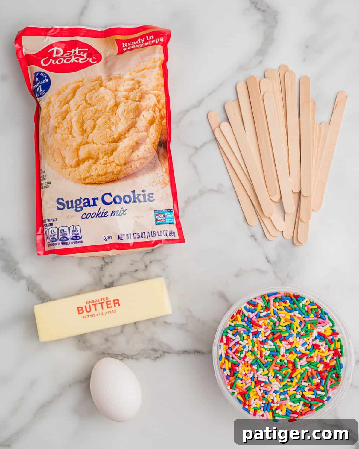 Cookie Pops: Make Handheld Cookies on a Stick 4 Ingredients on a marble counter: Betty Crocker sugar cookie mix, wooden popsicle sticks, a stick of unsalted butter, an egg, and a bowl of rainbow sprinkles.