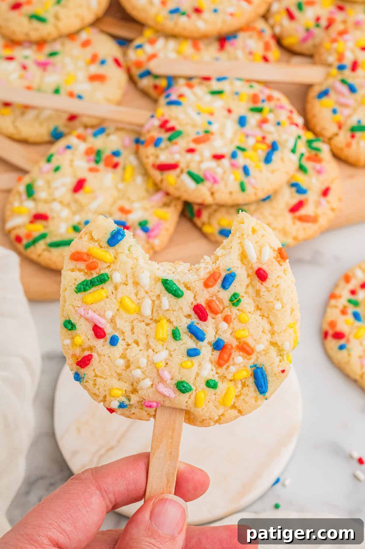 Cookie Pops: Make Handheld Cookies on a Stick 6 A hand holding a sprinkle sugar cookie on a stick with a bite taken out, more cookies on a wooden board behind.