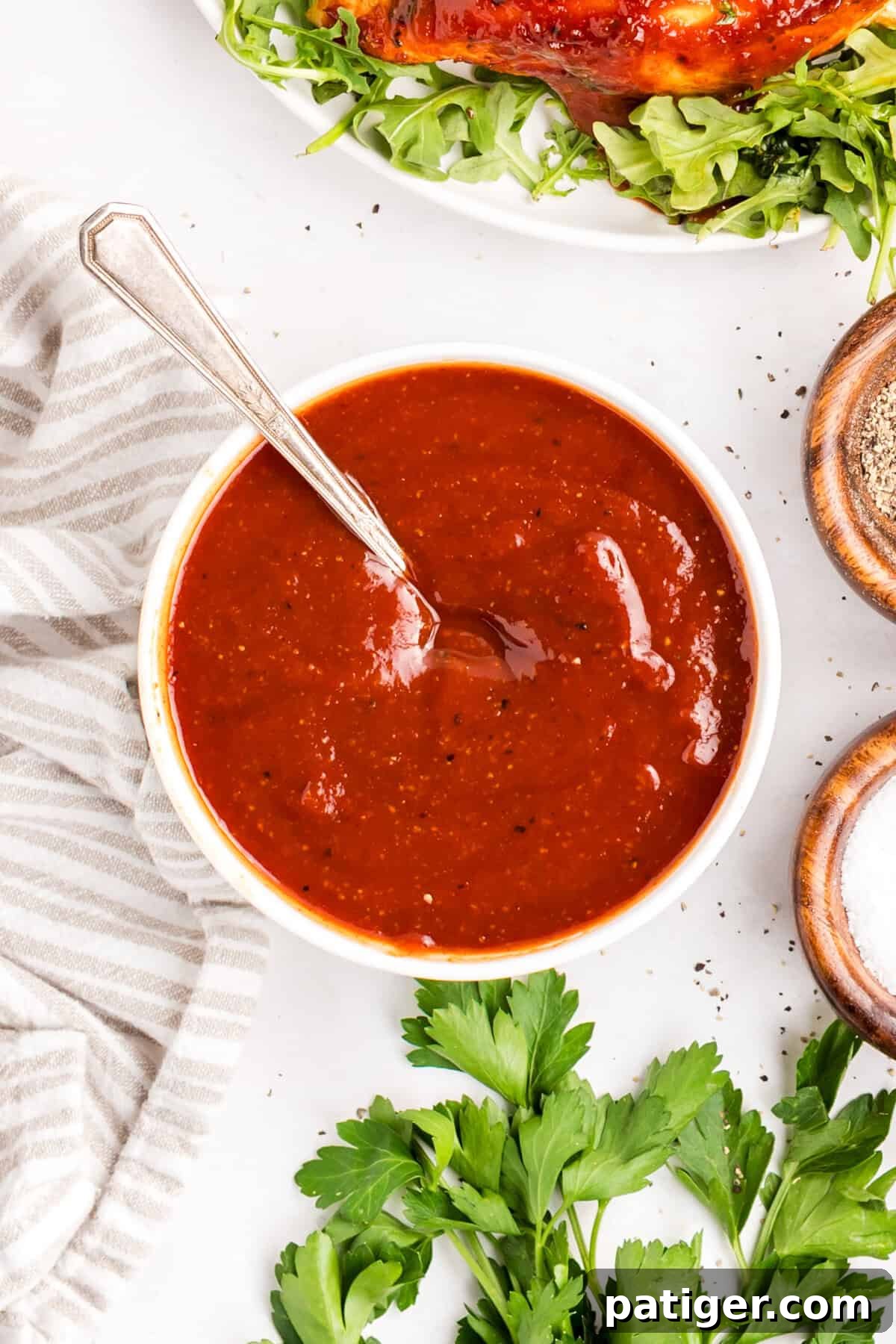 How to Make Tangy Homemade BBQ Sauce 2 Overhead bowl of homemade barbecue sauce with a spoon, a plate of sauced chicken and small dishes of salt and pepper alongside.