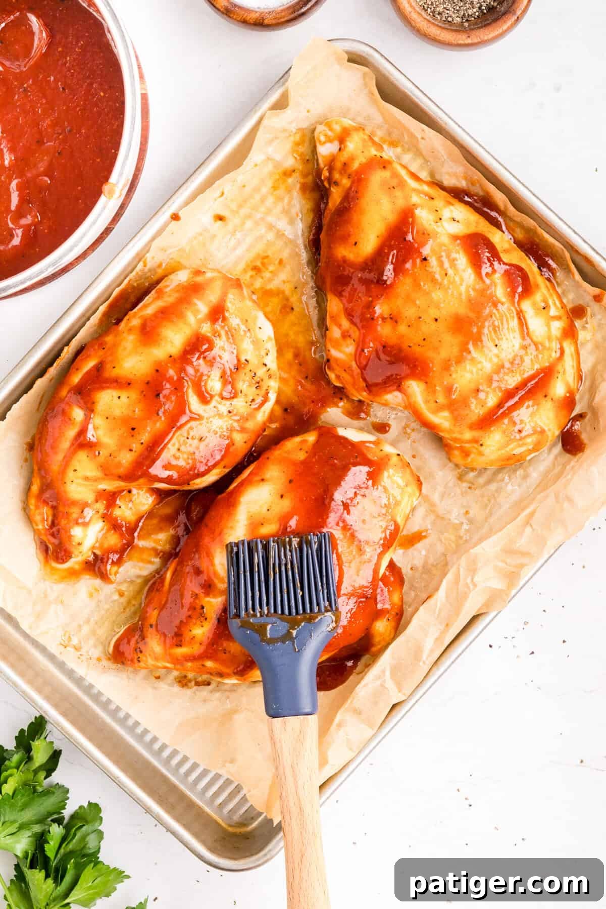 How to Make Tangy Homemade BBQ Sauce 5 A pastry brush spreading barbecue sauce over three raw chicken breasts on a parchment-lined sheet pan.