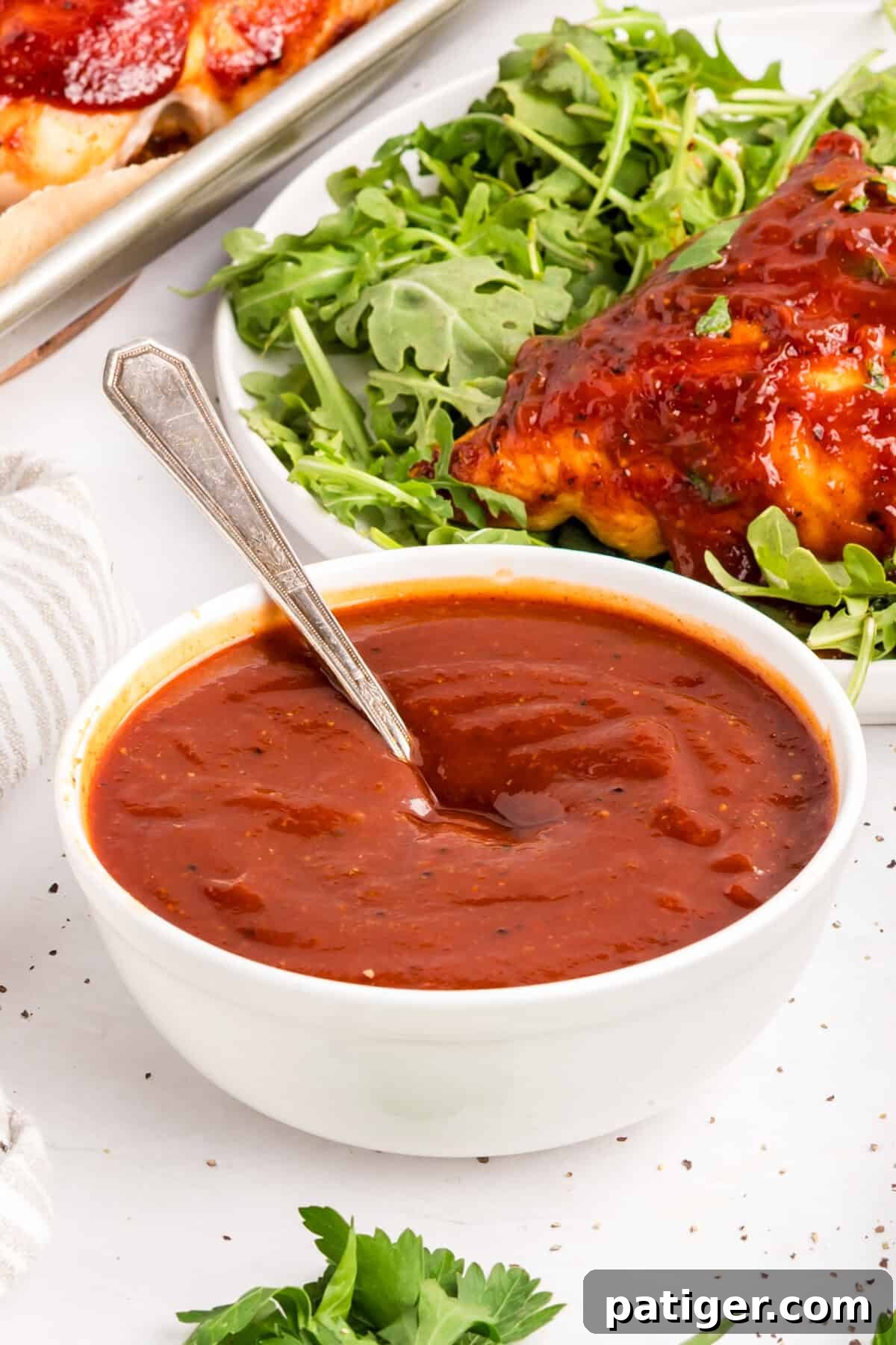 How to Make Tangy Homemade BBQ Sauce 6 Homemade BBQ sauce in a white bowl with a spoon, a plate of BBQ chicken over arugula behind it.