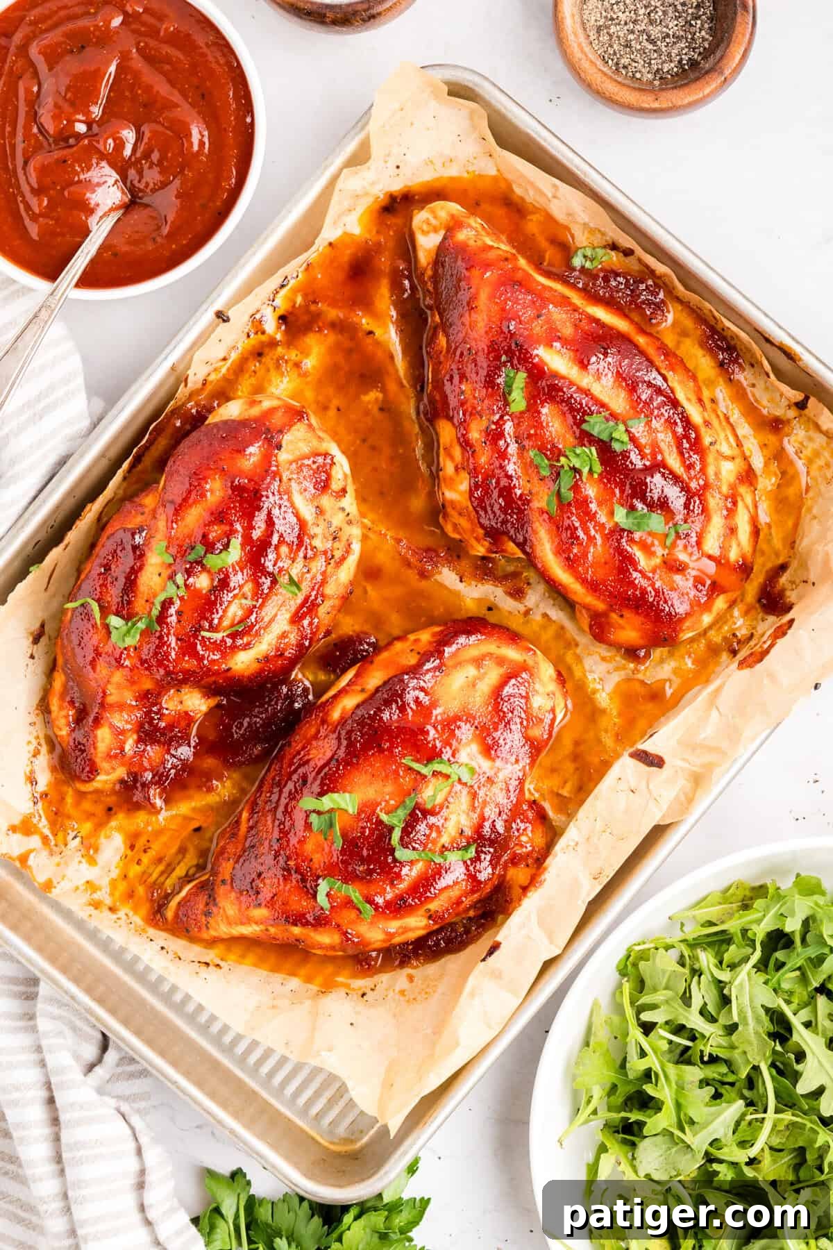 Oven-Baked BBQ Chicken Breasts with Sticky Glaze 2 Overhead of baked BBQ chicken breast on a parchment-lined sheet pan with a bowl of BBQ sauce.
