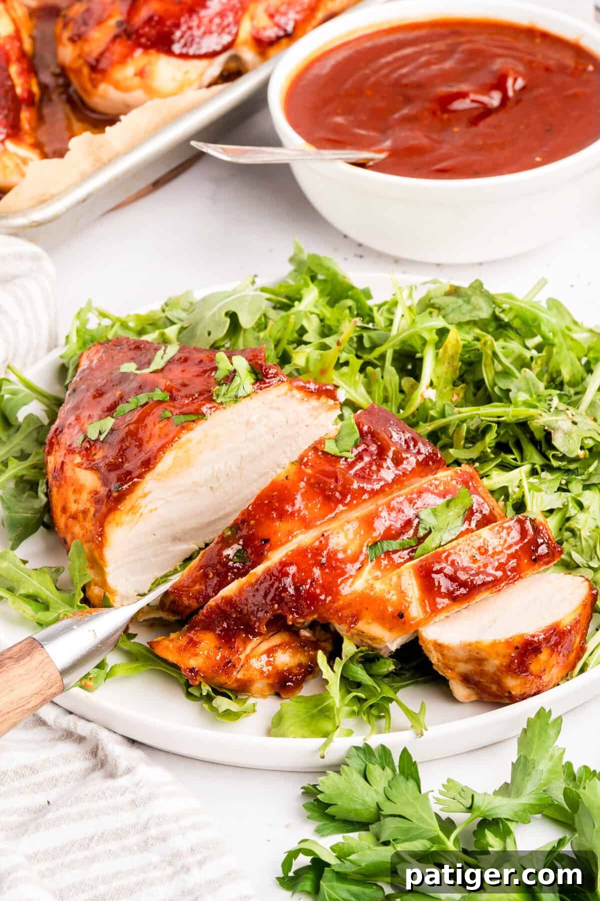 Oven-Baked BBQ Chicken Breasts with Sticky Glaze 6 Sliced baked BBQ chicken breast on a bed of arugula with a knife and a bowl of sauce behind it.