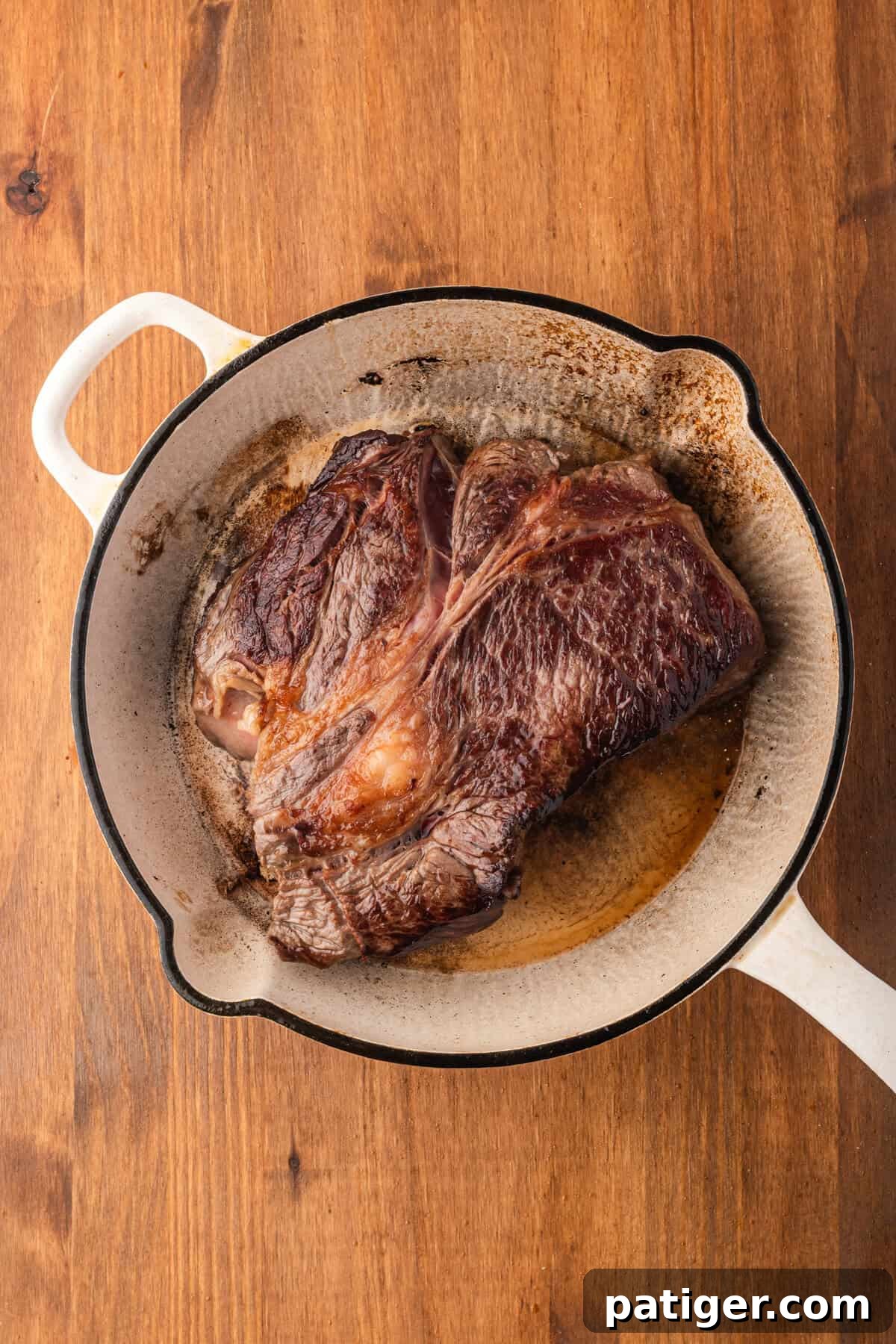 Top-down view of a seared chuck roast in a white Dutch oven on a wood surface.