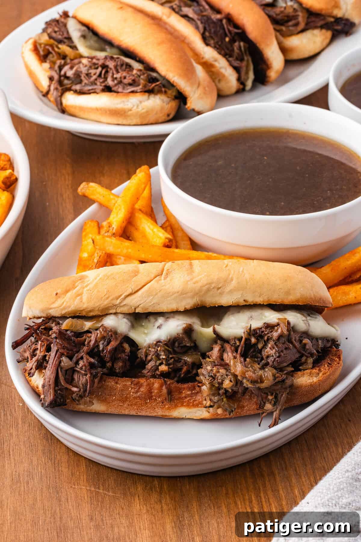 A French dip sandwich plated next to a small bowl of au jus and fries, with more sandwiches on a plate behind.
