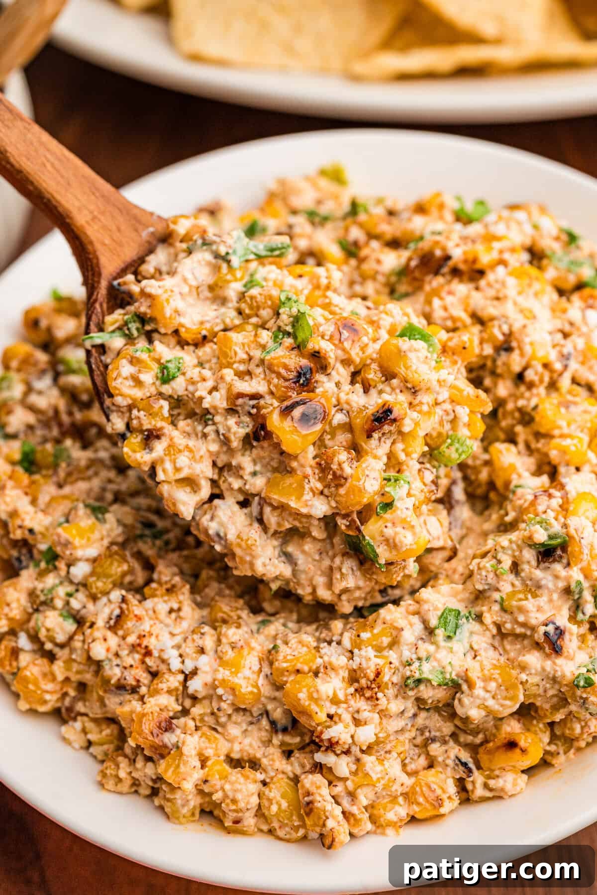Creamy Mexican Street Corn Dip with Cotija and Lime 7 A wooden spoon lifting a scoop of creamy corn dip from a white bowl, tortilla chips in the background.