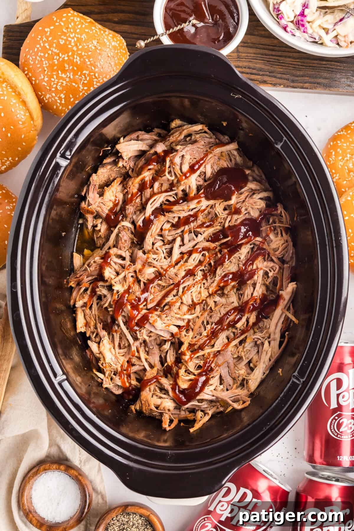 Overhead of a slow cooker full of shredded pork drizzled with barbecue sauce, sesame buns and Dr Pepper cans nearby.