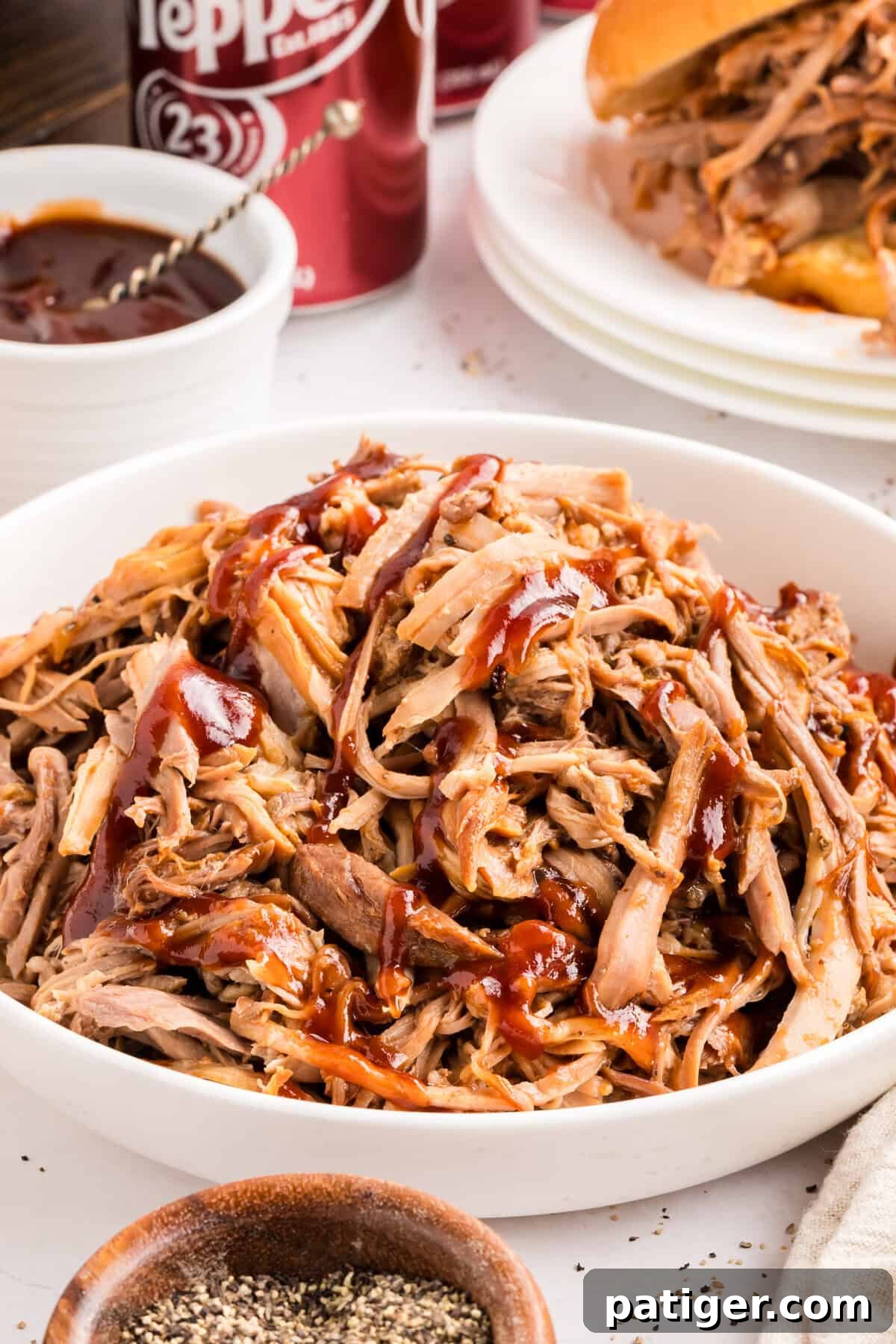Dr Pepper pulled pork in a white bowl drizzled with barbecue sauce, a Dr Pepper can and a sandwich behind it.
