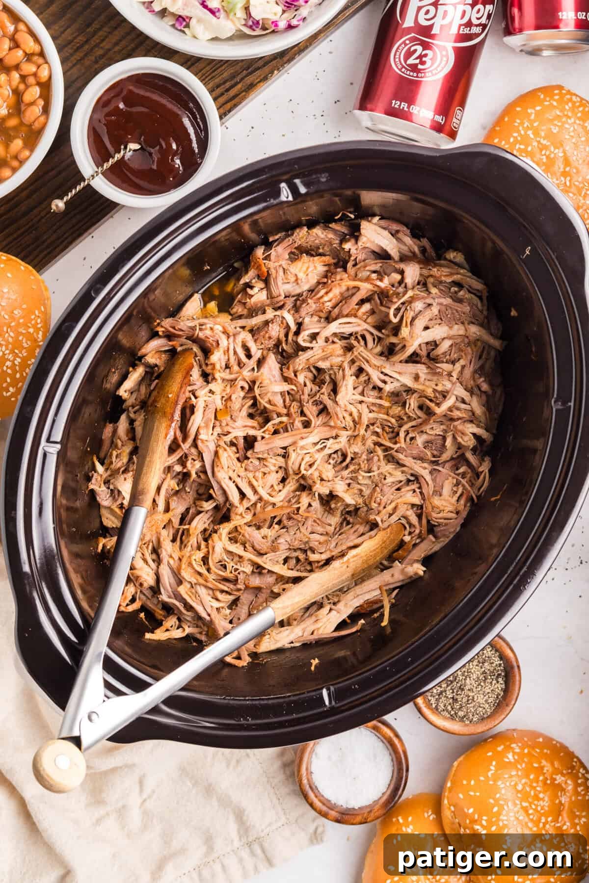 Overhead of shredded pork in a black slow cooker with wooden tongs, surrounded by buns, baked beans, coleslaw, and Dr Pepper.
