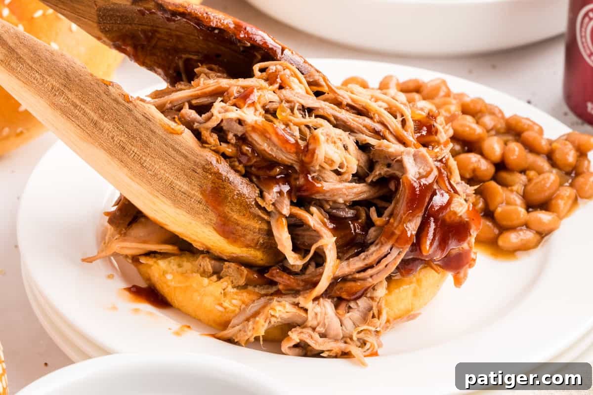 Wooden tongs piling barbecue pulled pork onto a sesame bun with baked beans on a white plate.