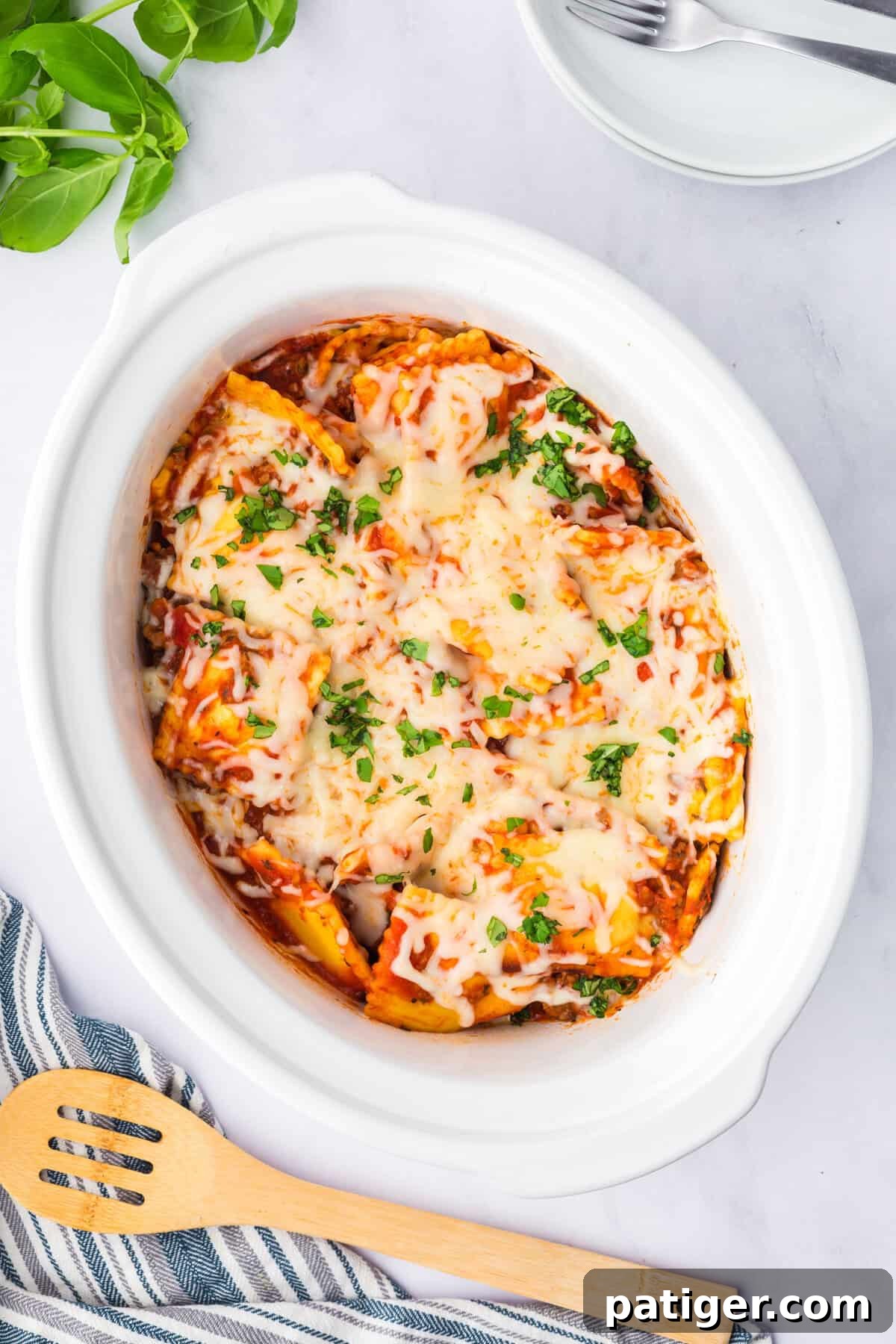 Overhead view of a wooden spoon in a slow cooker full of cheesy ravioli topped with basil.
