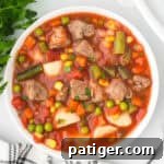 Square overhead view of a white bowl filled with beef, potatoes, green beans, corn and peas in tomato broth.