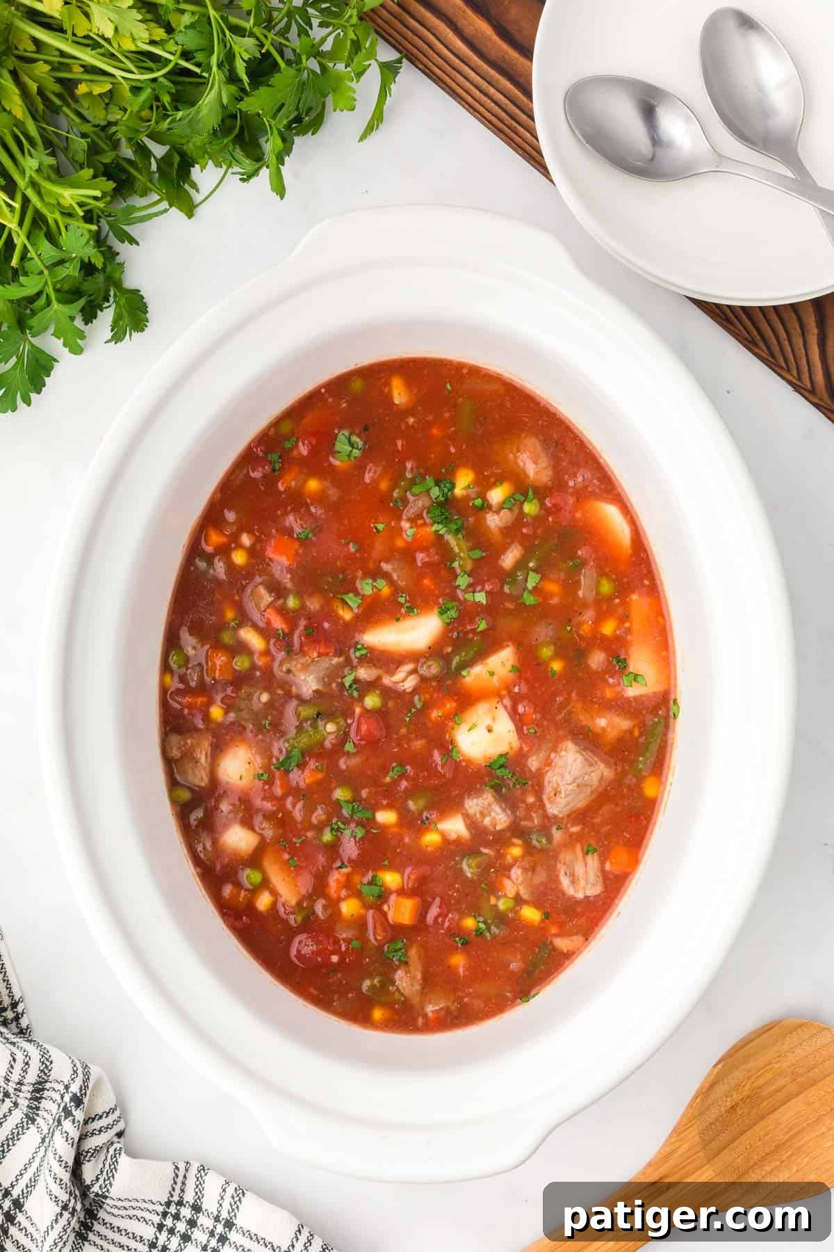Overhead view of slow cooker vegetable beef soup in a white crockpot with parsley, bowls and spoons alongside.