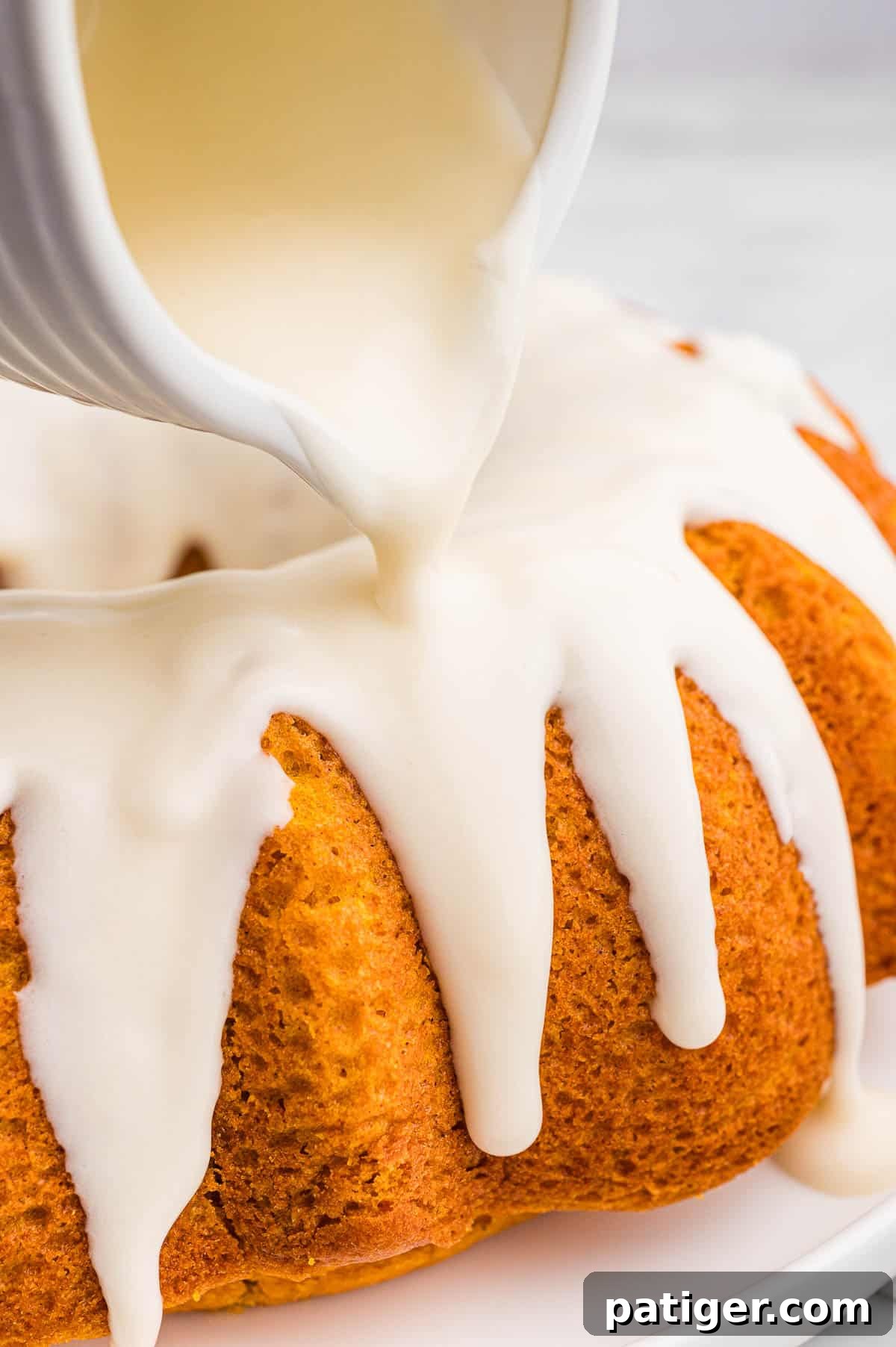 Moist Pumpkin Bundt Cake Made with Cake Mix 9 Cream cheese glaze being poured from a white pitcher over a pumpkin bundt cake and dripping down the ridges.