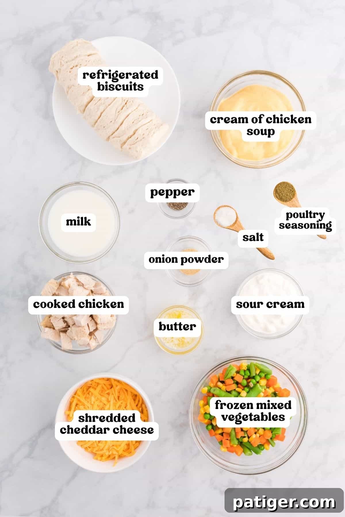One-Pan Chicken Pot Pie Casserole with Biscuits 4 Labeled bowls of cooked chicken, cheddar, frozen mixed vegetables, cream of chicken soup, sour cream, milk, butter, onion powder, poultry seasoning, salt, pepper, and refrigerated biscuits on marble.
