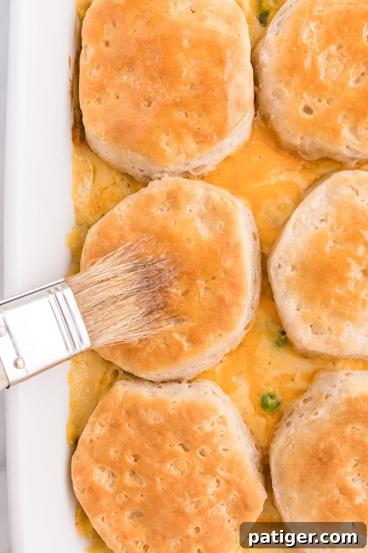 One-Pan Chicken Pot Pie Casserole with Biscuits 6 A pastry brush spreading melted butter over golden baked biscuits in a white casserole dish.