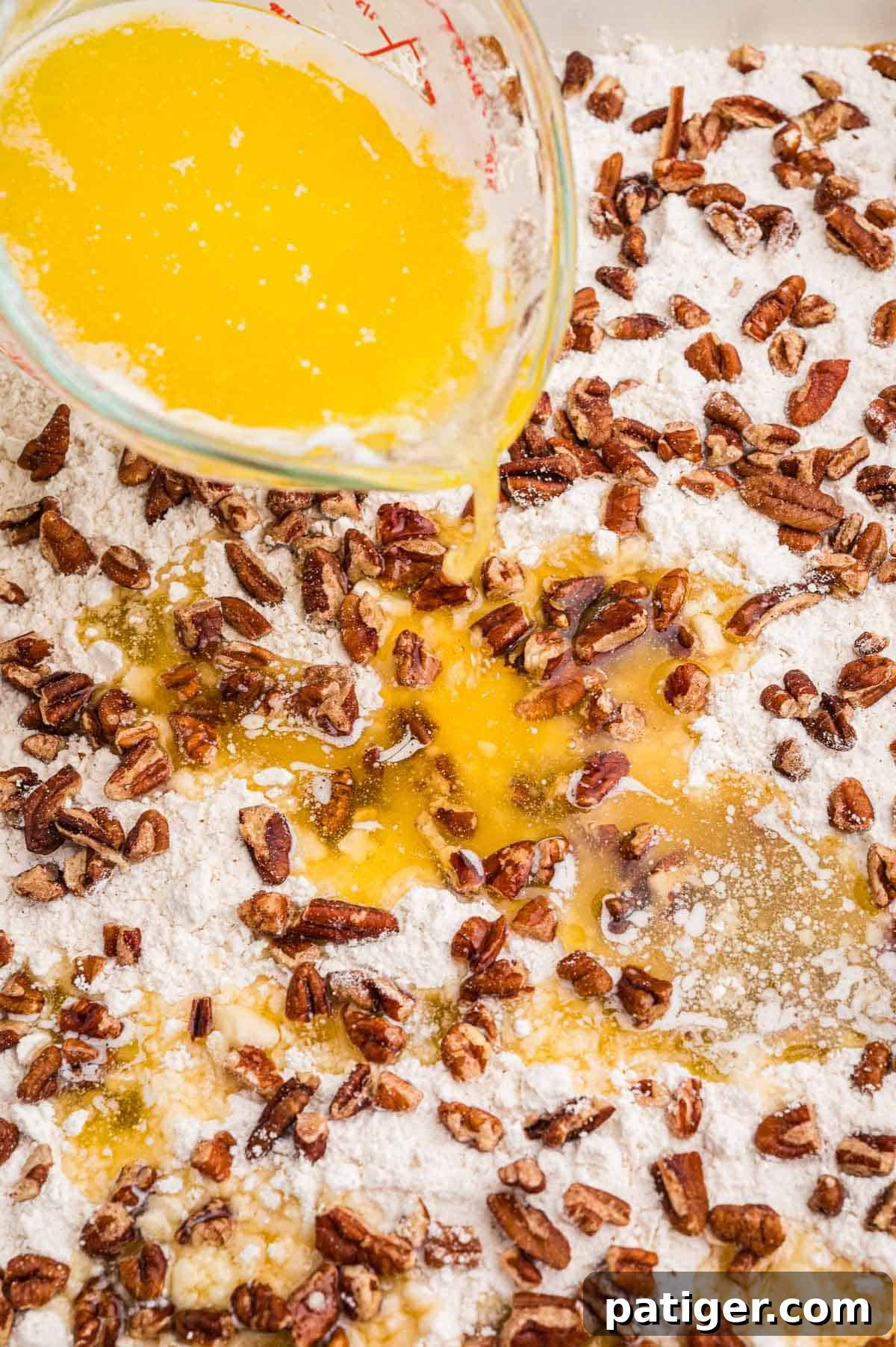Melted butter being poured from a glass measuring cup over dry cake mix and chopped pecans in a baking dish.