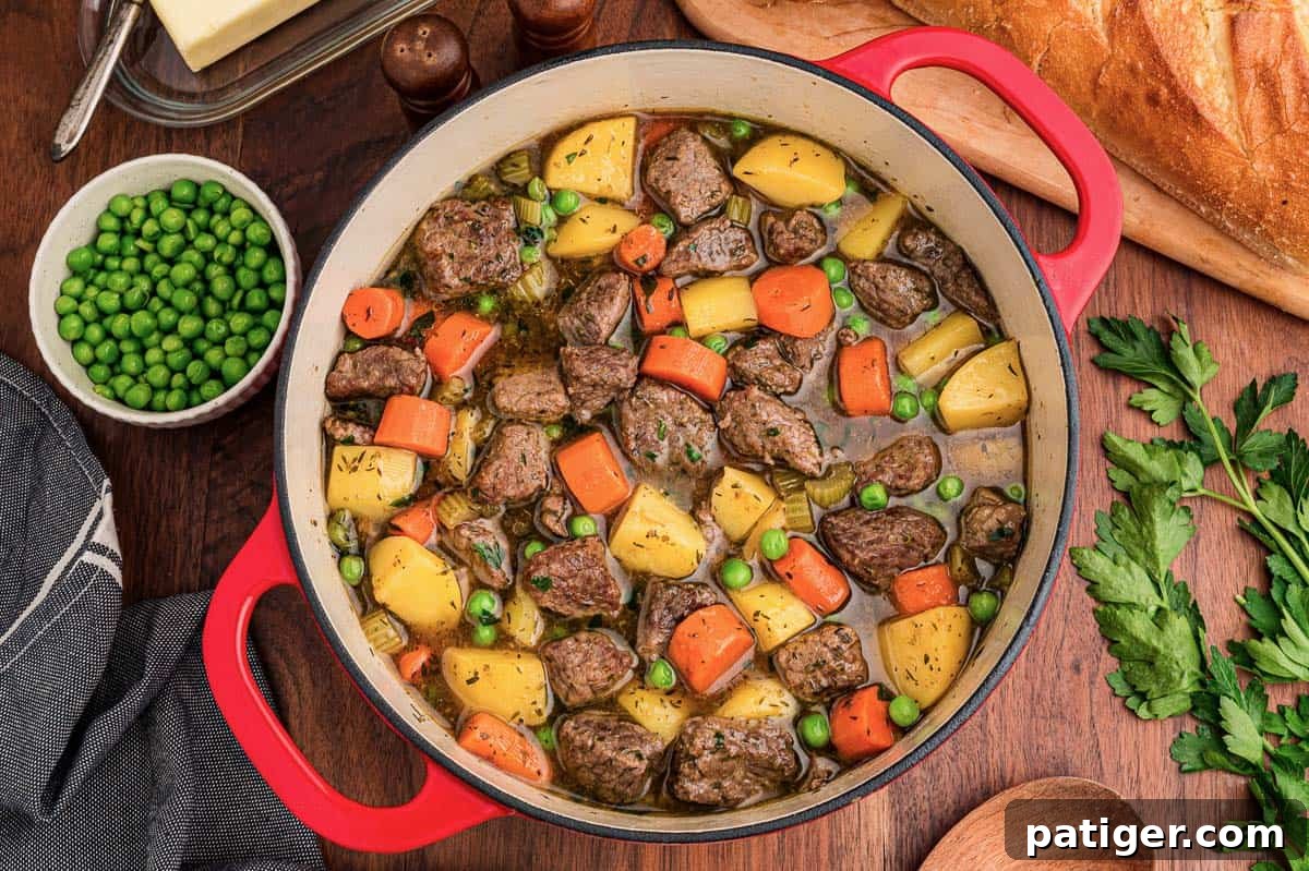 Classic Hearty Beef Stew Recipe with Tender Braised Beef 2 Easy beef stew with potatoes, carrots, celery, and peas in a red Dutch oven beside crusty bread.