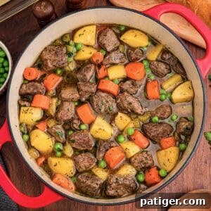 Overhead view of a red Dutch oven filled with beef, potatoes, carrots, celery, and peas.