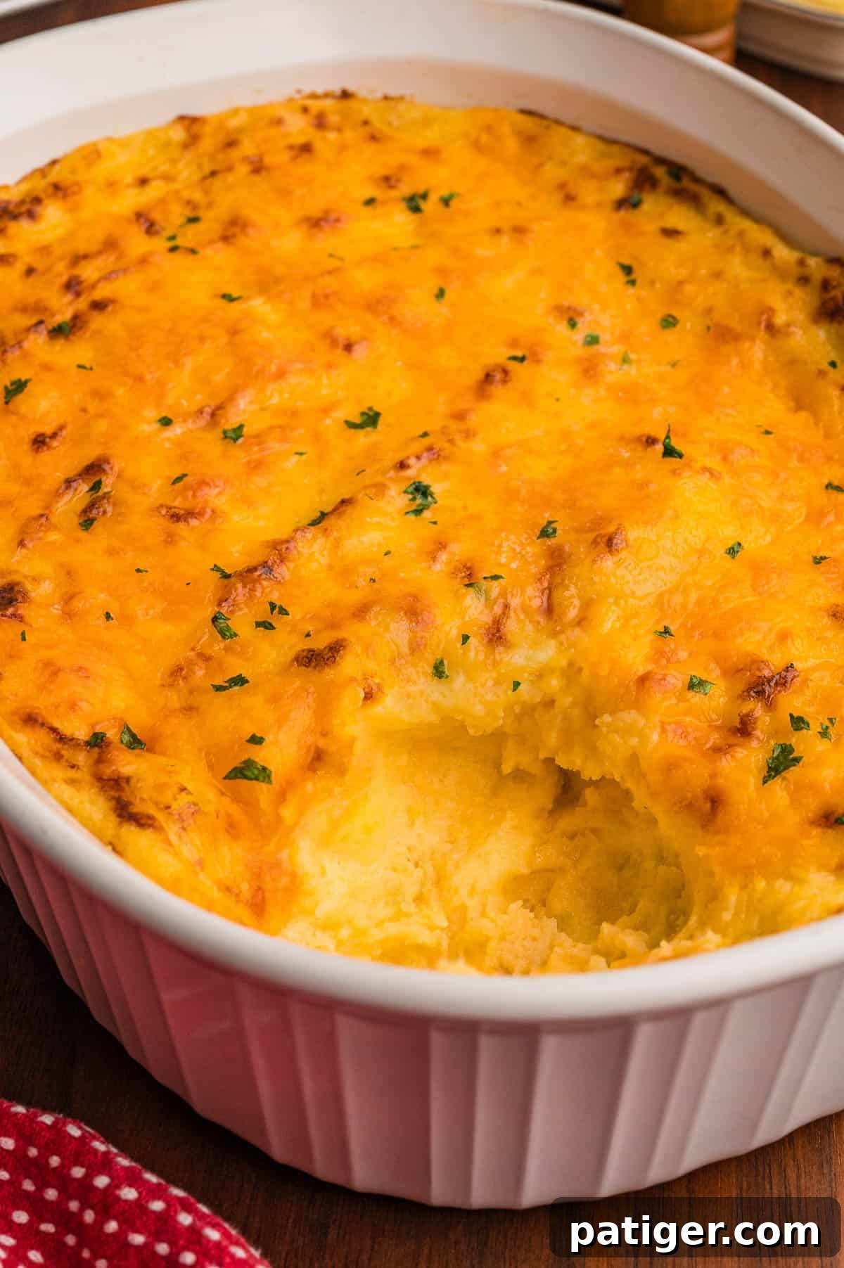 White oval baking dish of mashed potatoes with a golden cheddar crust and a scoop removed, next to a red polka dot towel.