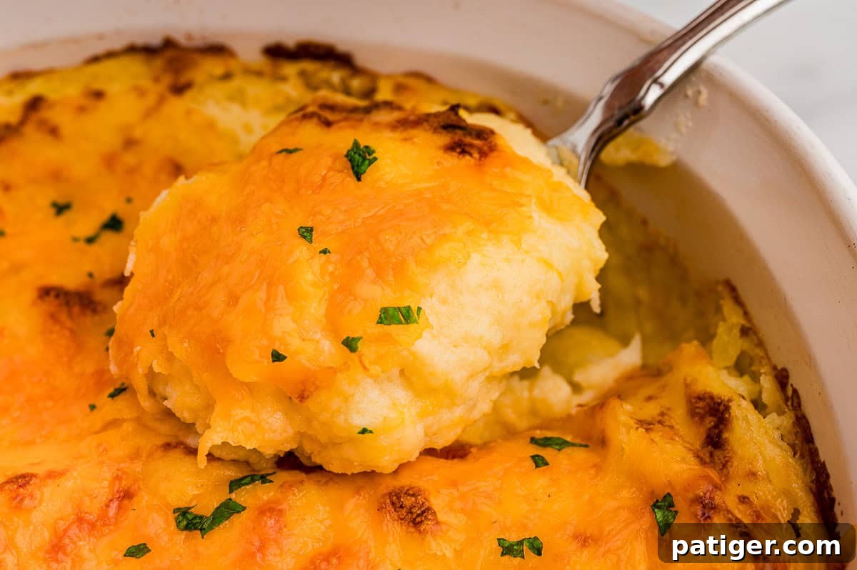 Close-up of a serving spoon scooping creamy potatoes topped with melted, browned cheddar and chopped parsley.