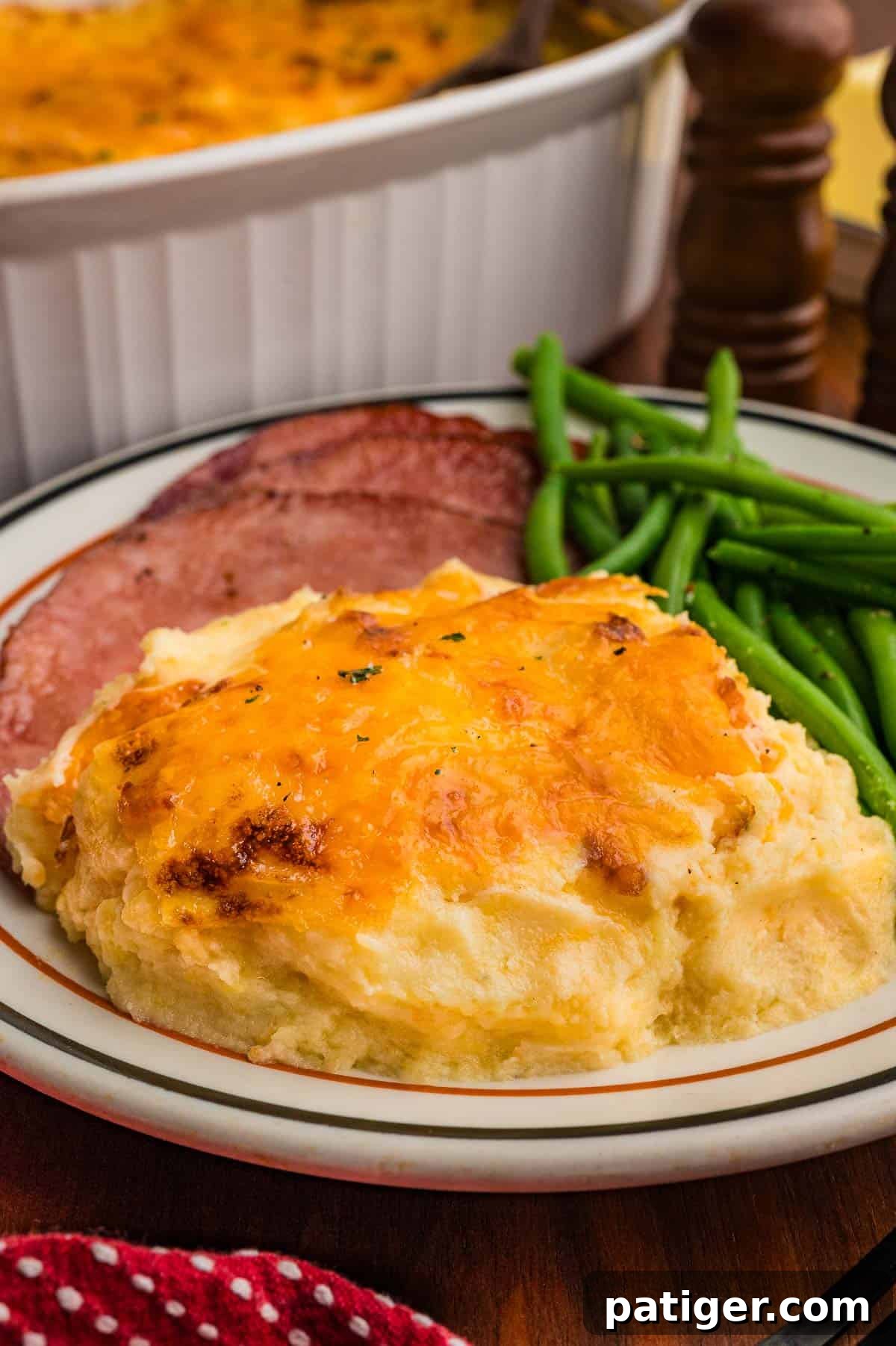 A serving of cheddar-topped mashed potatoes on a plate with sliced ham and green beans, with the casserole dish behind it.