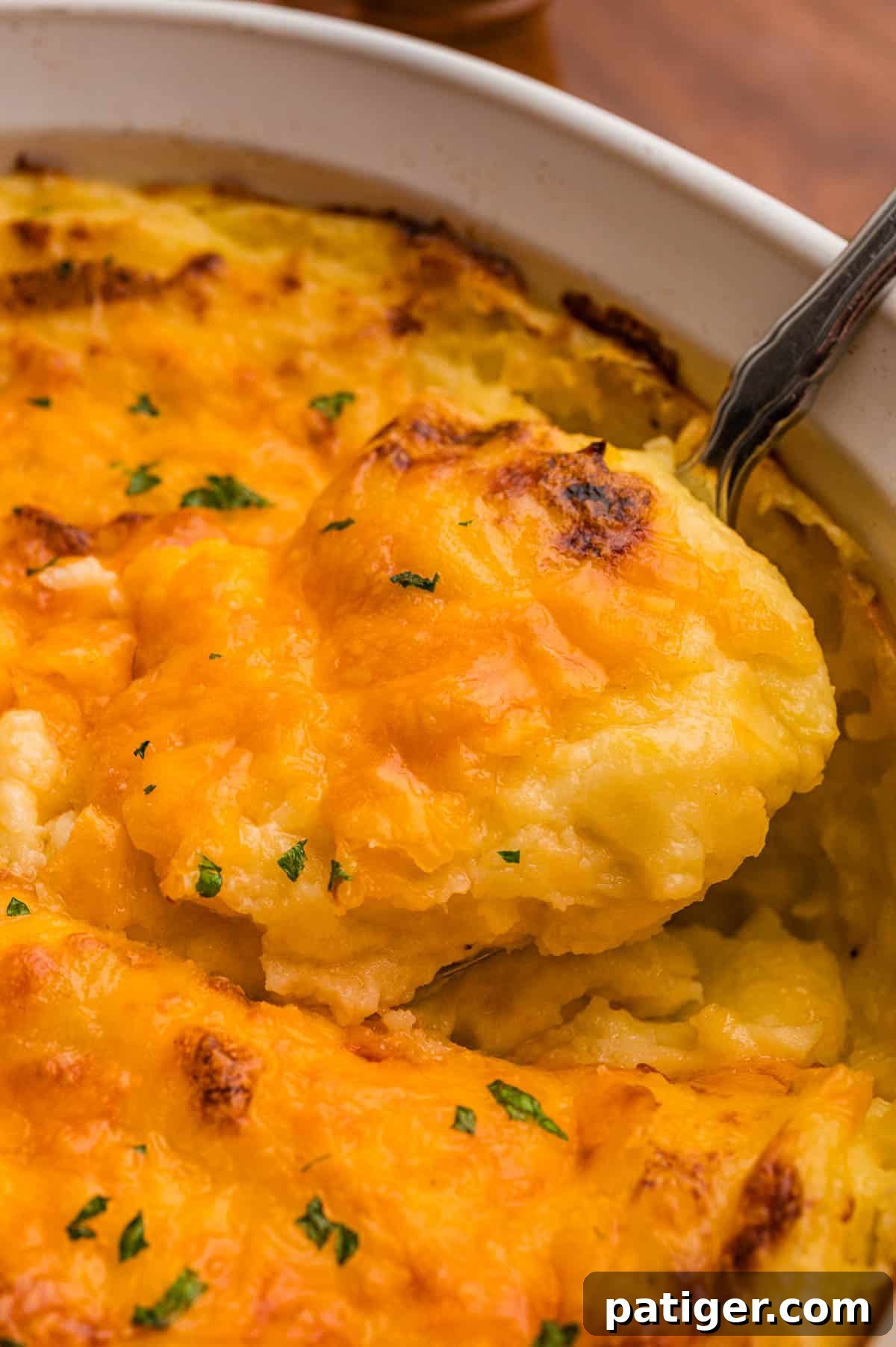 A spoon lifting a scoop of mashed potatoes with a melted, browned cheddar top from a white dish.
