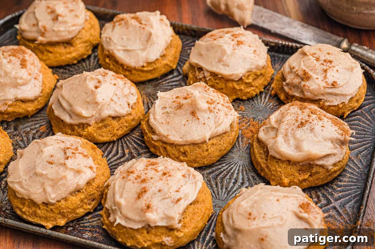 Frosted pumpkin cookies dusted with cinnamon on a dark metal pan with a frosting knife behind.