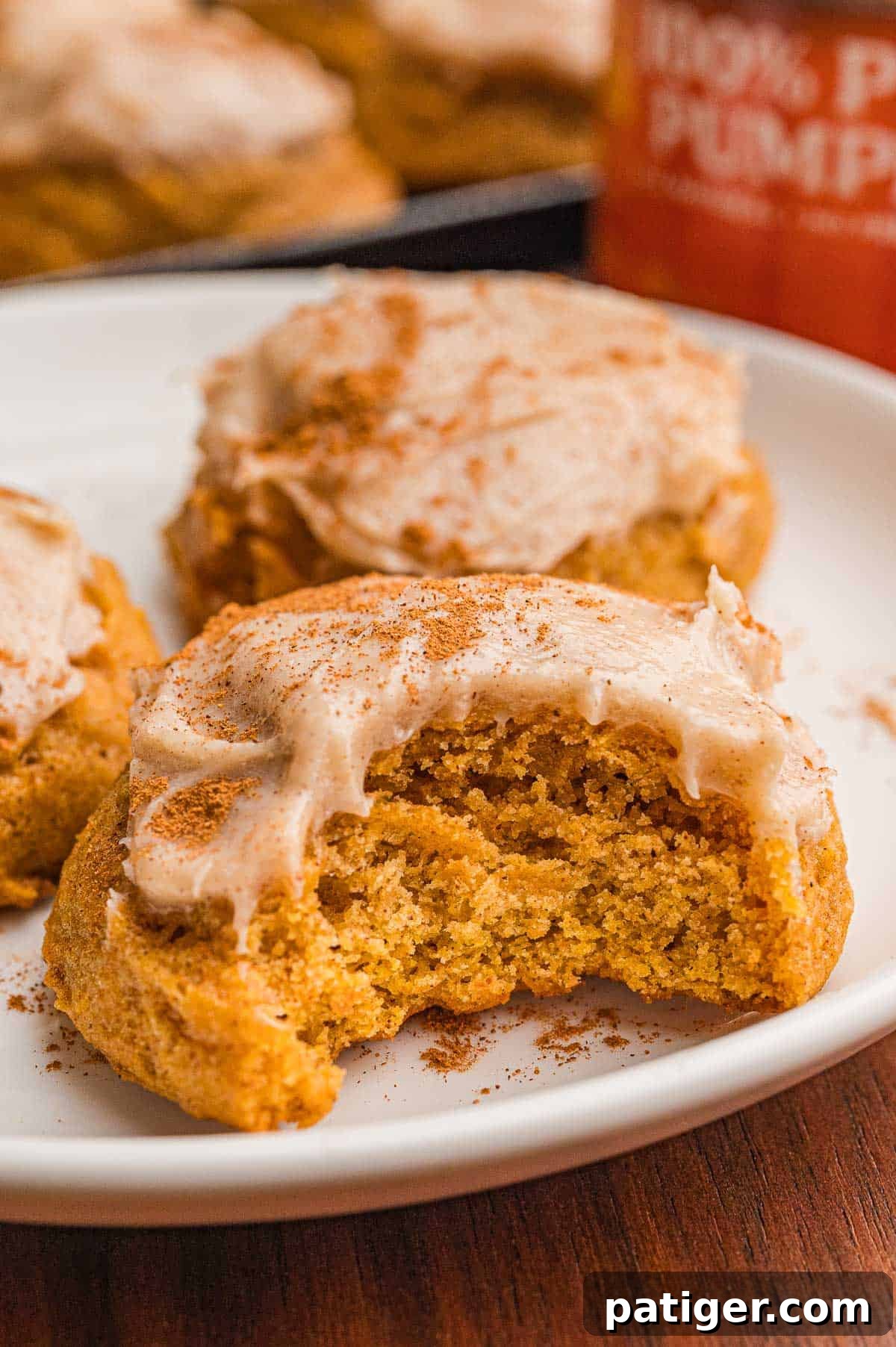 A frosted pumpkin cookie with a bite taken on a white plate, dusted with cinnamon.