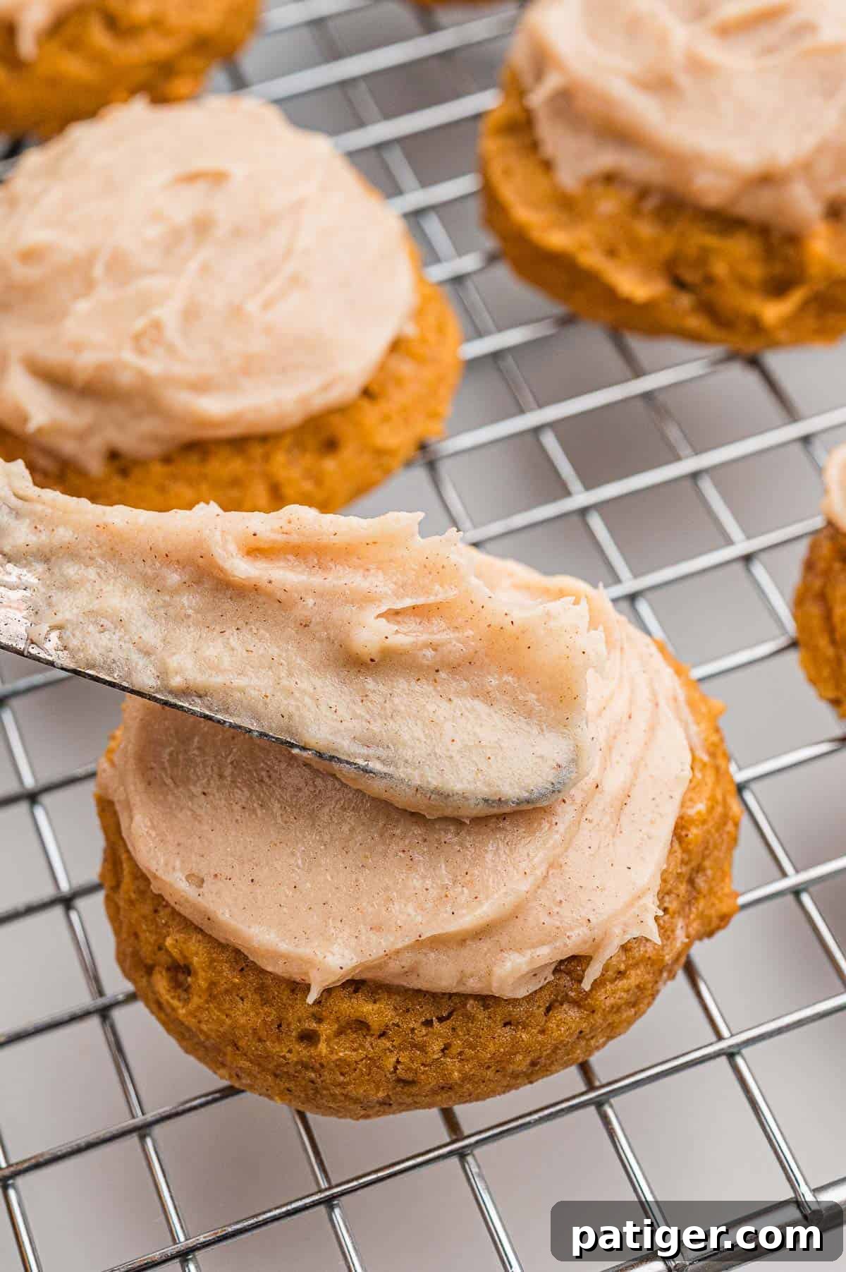 An offset spatula spreading cinnamon cream cheese frosting onto a cookie on a wire cooling rack.