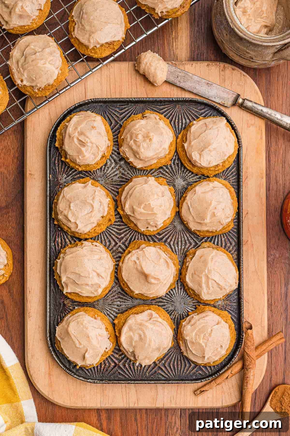 Overhead view of twelve frosted pumpkin cookies in a metal pan on a wooden board with cinnamon sticks.