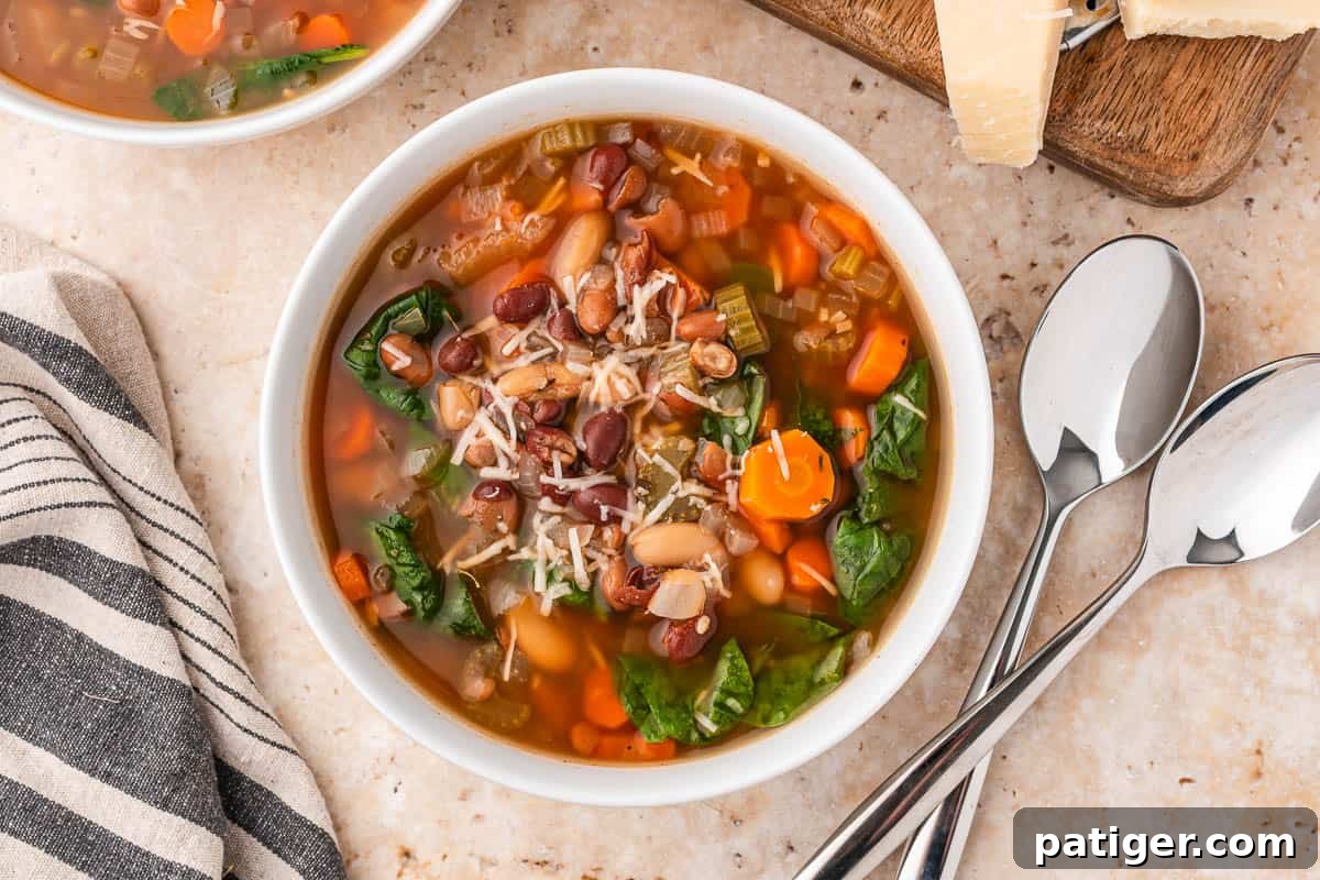 Overhead view of a bowl of bean soup with Parmesan, two spoons, and a striped towel beside it.