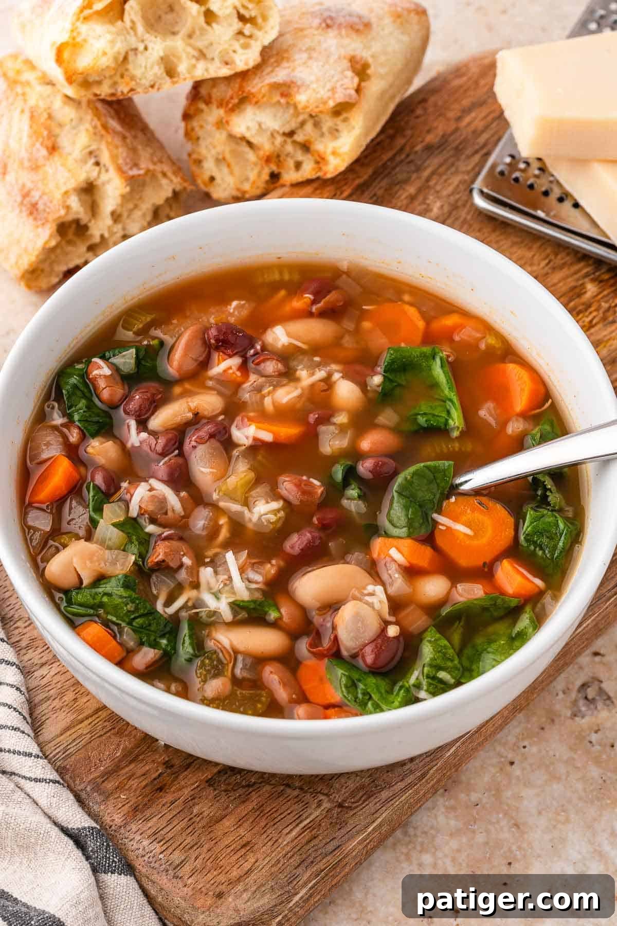 Bowl of three bean soup with carrots and spinach on a wooden board, with bread and a Parmesan grater behind it.