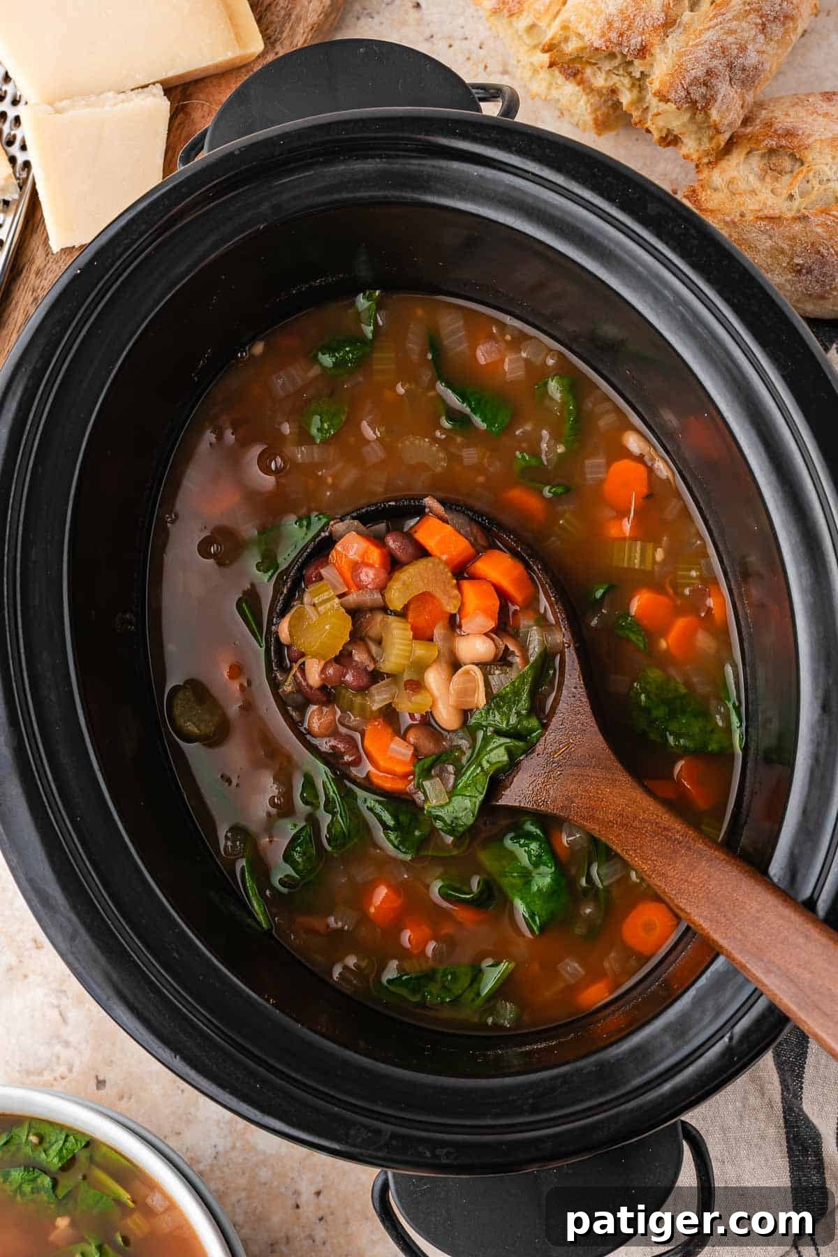 Overhead view of a black slow cooker of bean soup with a ladle, with crusty bread and Parmesan alongside.