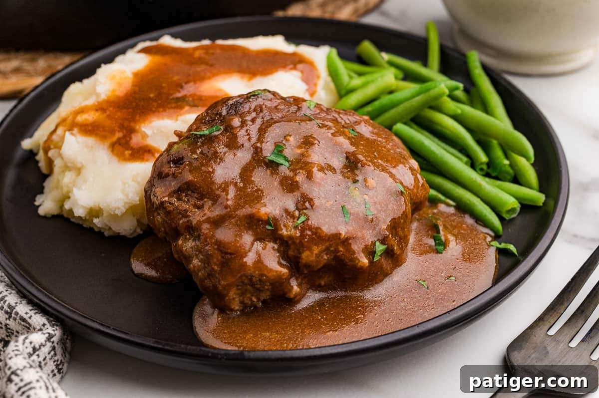 Salisbury steak recipe served on a black plate with mashed potatoes, brown gravy, and green beans.