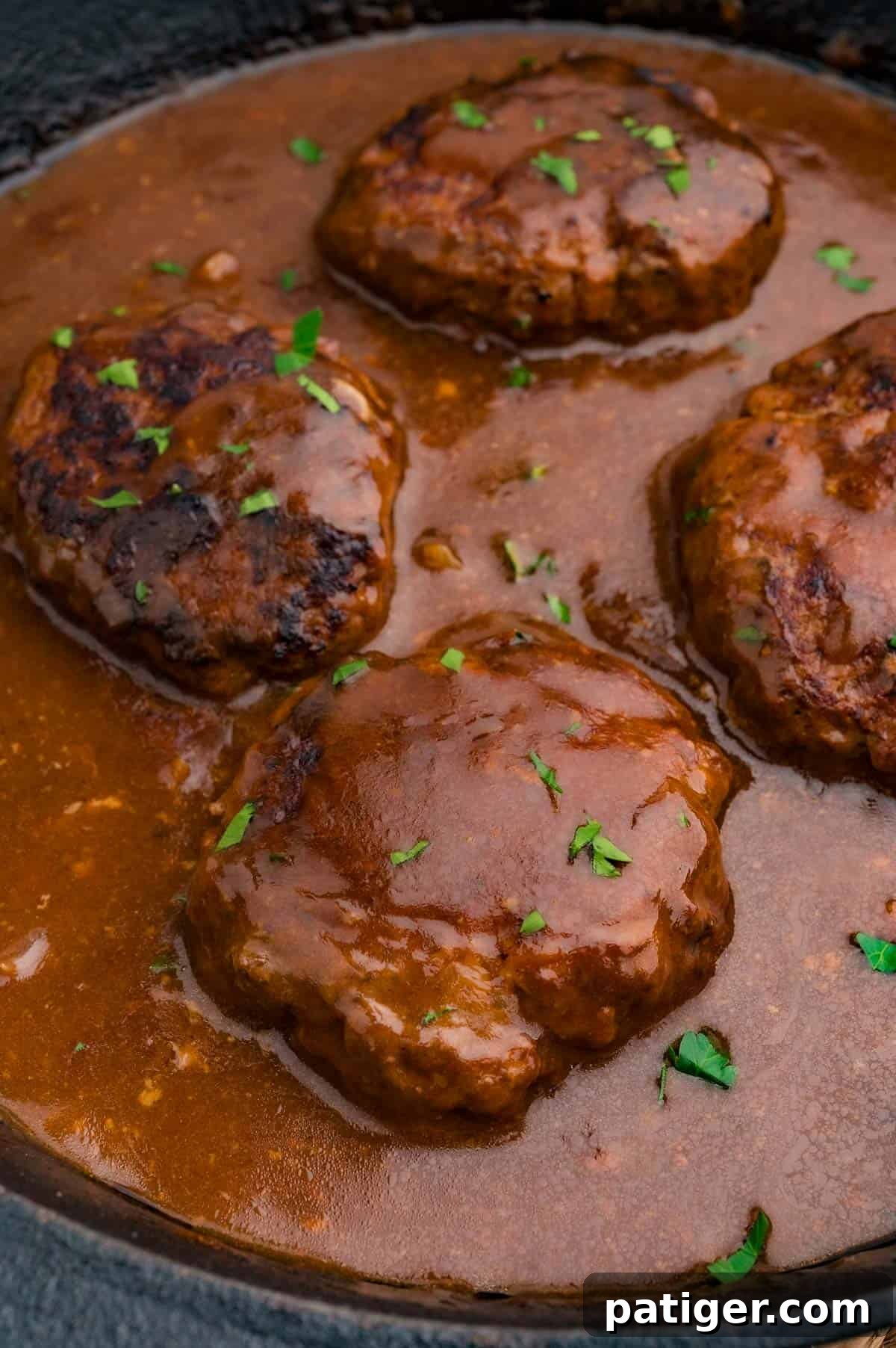 Four browned beef patties simmering in brown gravy in a cast iron skillet, sprinkled with parsley.