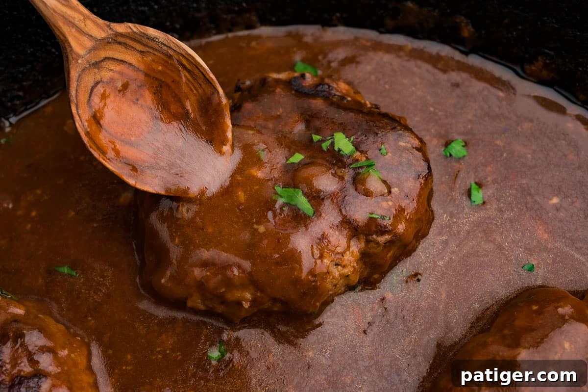 A wooden spoon pouring brown gravy over a seared beef patty in a skillet.