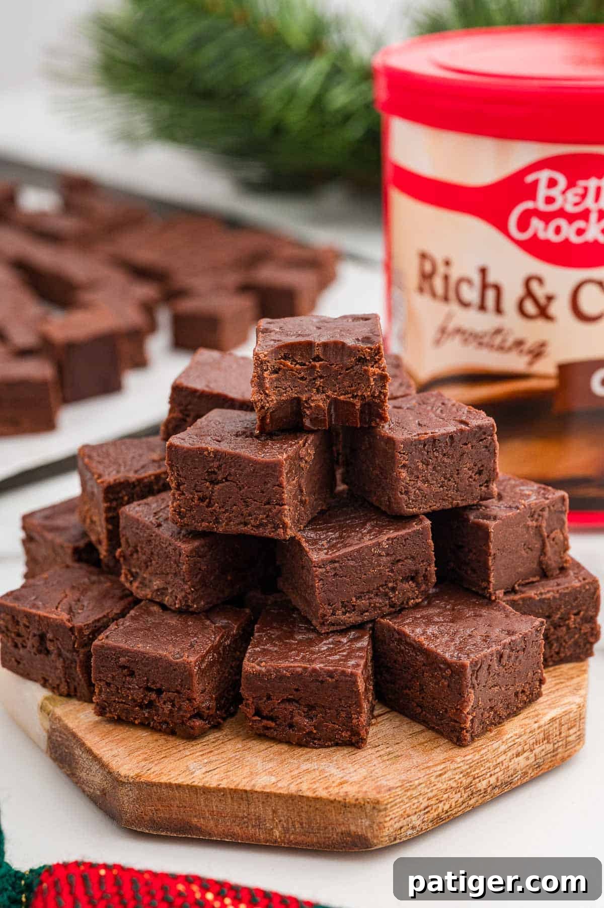Two-Ingredient Chocolate Fudge Recipe 2 Pyramid of chocolate fudge squares stacked on a round wooden board with a tub of chocolate frosting behind it.