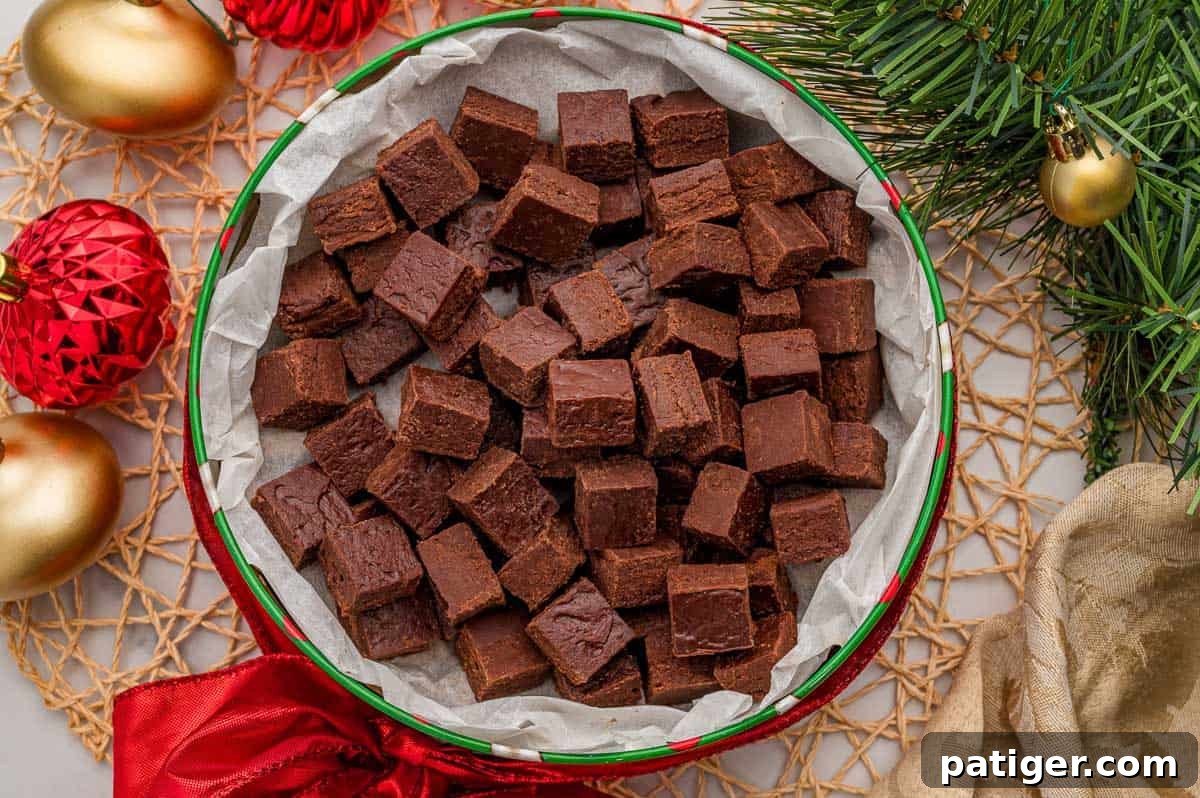Two-Ingredient Chocolate Fudge Recipe 3 Squares of 2 ingredient fudge with frosting piled in a round holiday tin lined with parchment paper, with red and gold ornaments and pine branches around it.