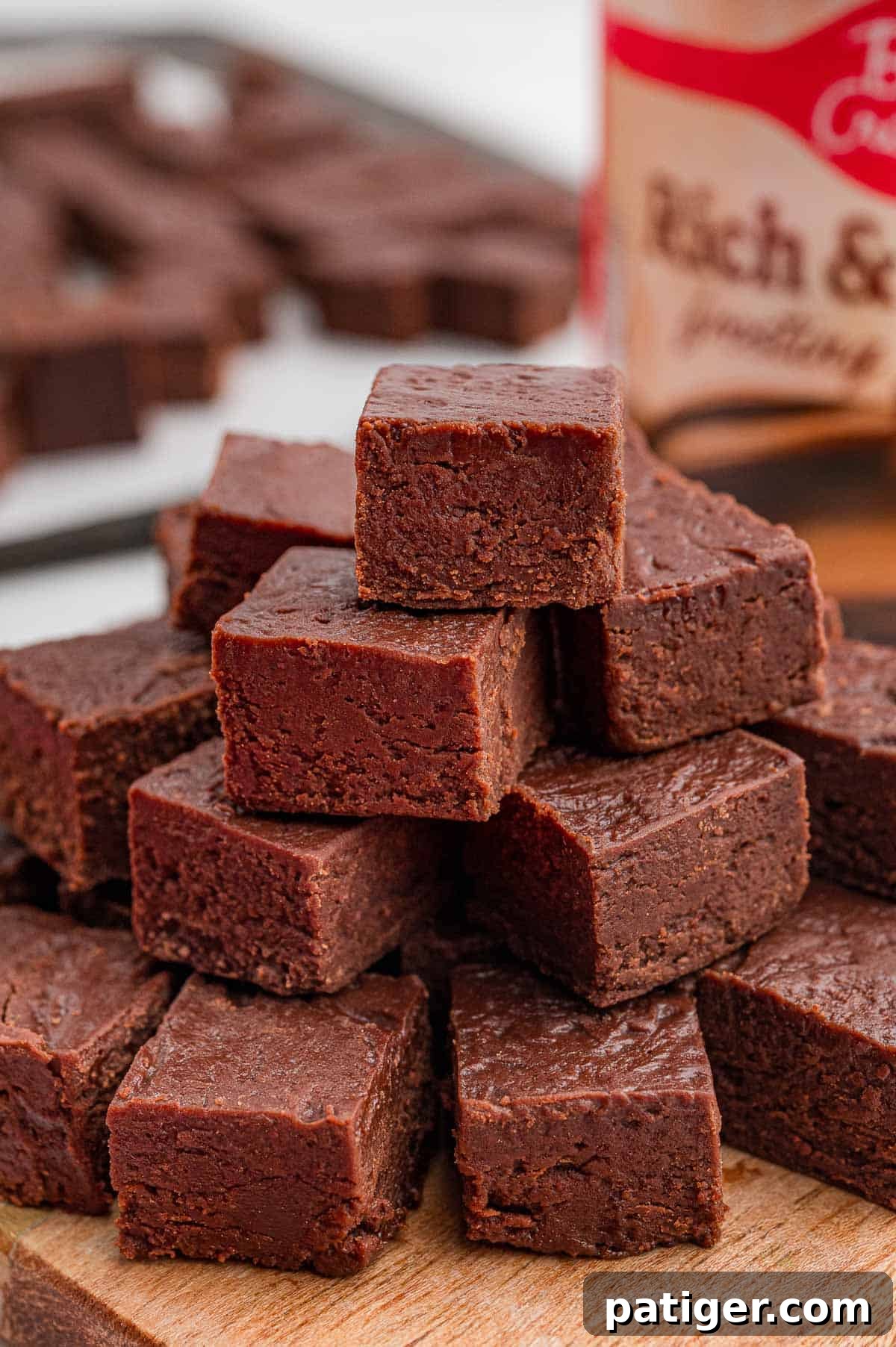Two-Ingredient Chocolate Fudge Recipe 7 Close-up of chocolate fudge squares stacked on a wooden board with a frosting tub behind them.