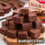 Two-Ingredient Chocolate Fudge Recipe 8 Fudge squares stacked on a wooden board with the top piece missing a bite and a frosting tub in the background.